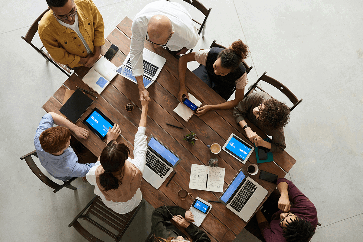 A group of eight people sit around a table. Two reach over to shake hands from either side.