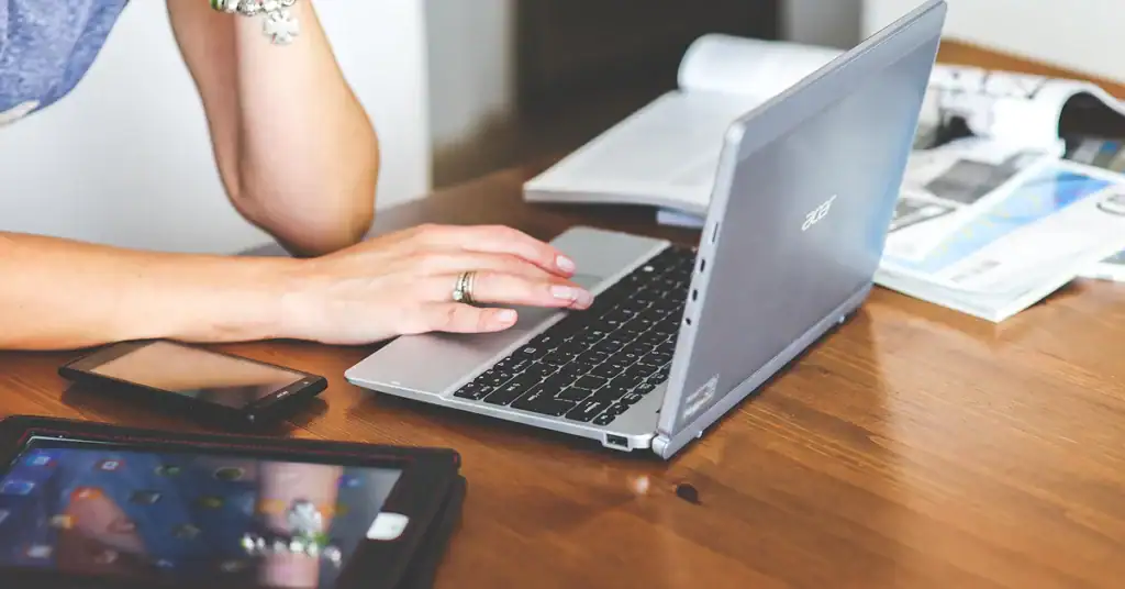 Person working on a laptop with smartphone and tablet on a wooden desk.